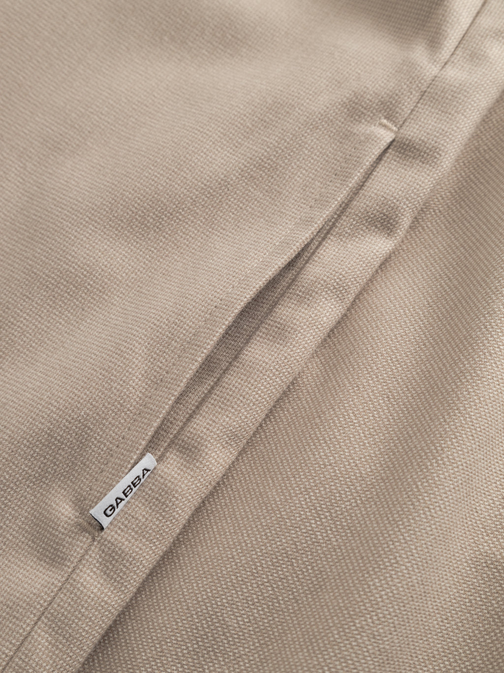 Close-up of the GABtogger Base Deme LS Overshirt - Birch by Gabba, showing textured beige fabric, a neat seam, and a white label near the edge that highlights the brand’s durable, precise stitch work.