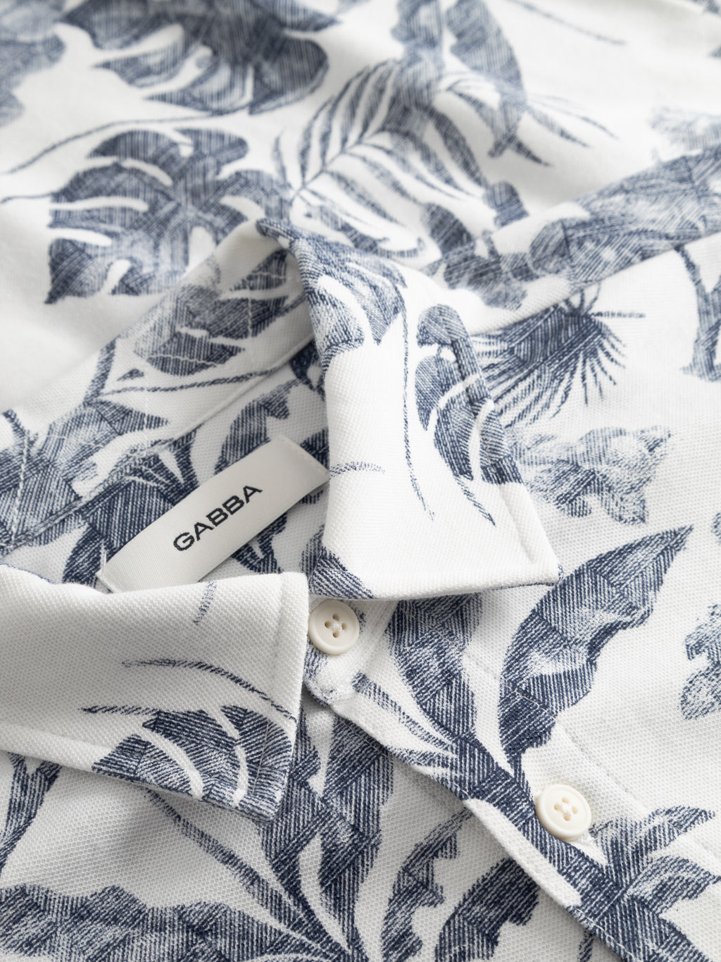Close-up of the GABsven Reg Leaves SS Shirt - Flower Mix by Gabba: a white short-sleeve shirt with blue tropical leaf prints in soft piqué jersey cotton, two white buttons, and a collar label featuring the Gabba brand name.
