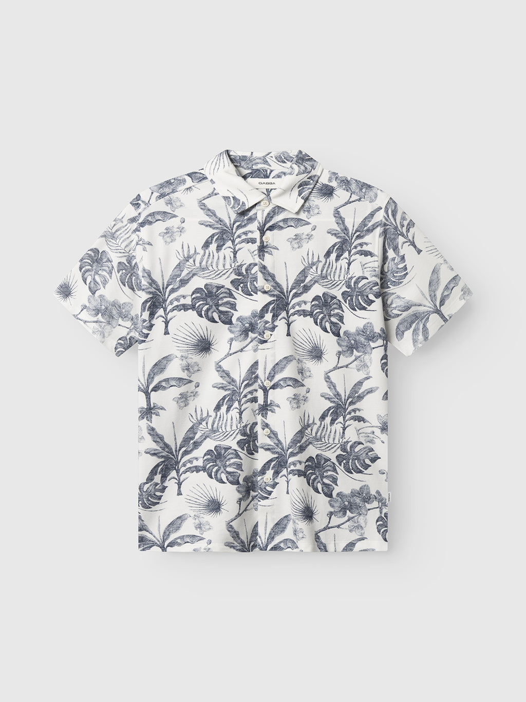 The Gabba GABsven Reg Leaves SS Shirt - Flower Mix is a regular-fit, short-sleeve button-up in piqué jersey cotton with a white base and navy tropical floral print, shown flat on a light gray background.