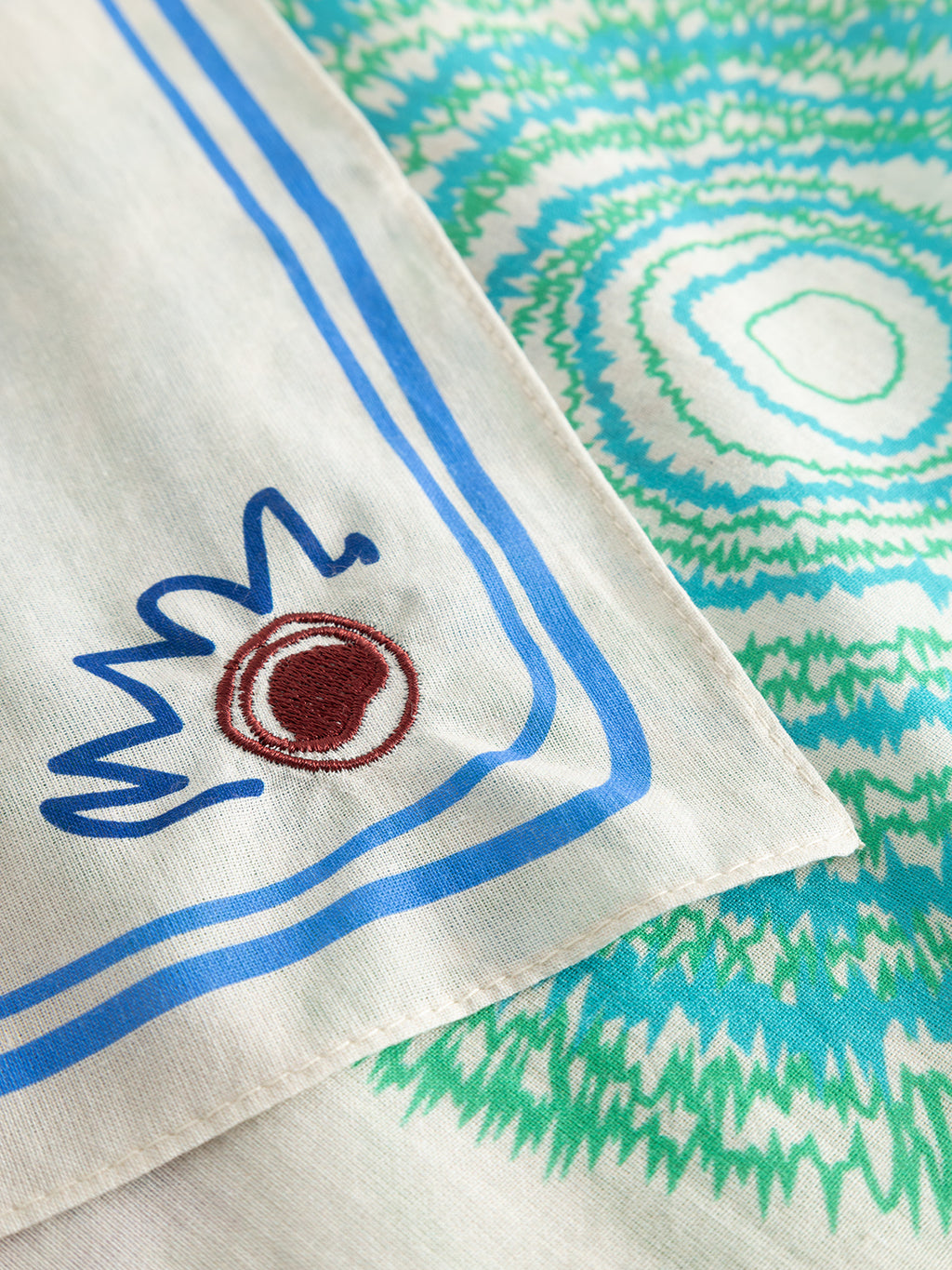 A close-up of the Gabba GABnando Bandana - Multi Pattern in a soft cotton blend, featuring blue and green geometric designs with a corner edged in blue lines and maroon-and-blue abstract accents.