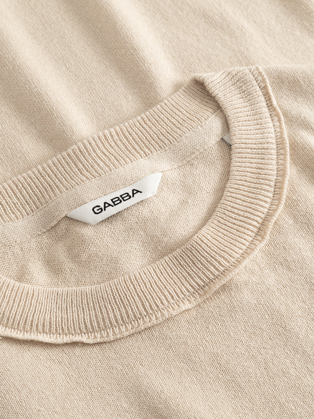 Close-up of the GABfumo Spy LS Knit - Sand by Gabba, a beige regular-fit cotton sweater featuring a flat-knit texture and visible stitching, with a white collar tag marked 