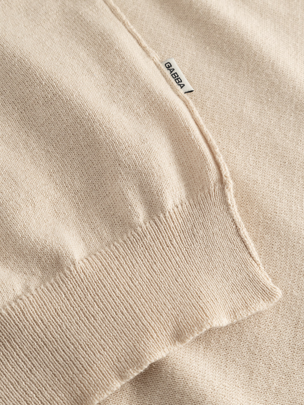 A close-up of the GABfumo Spy LS Knit - Sand by Gabba shows its beige regular-fit, soft cotton knit with a ribbed hem and a small white Gabba tag, highlighting the flat-knit construction.