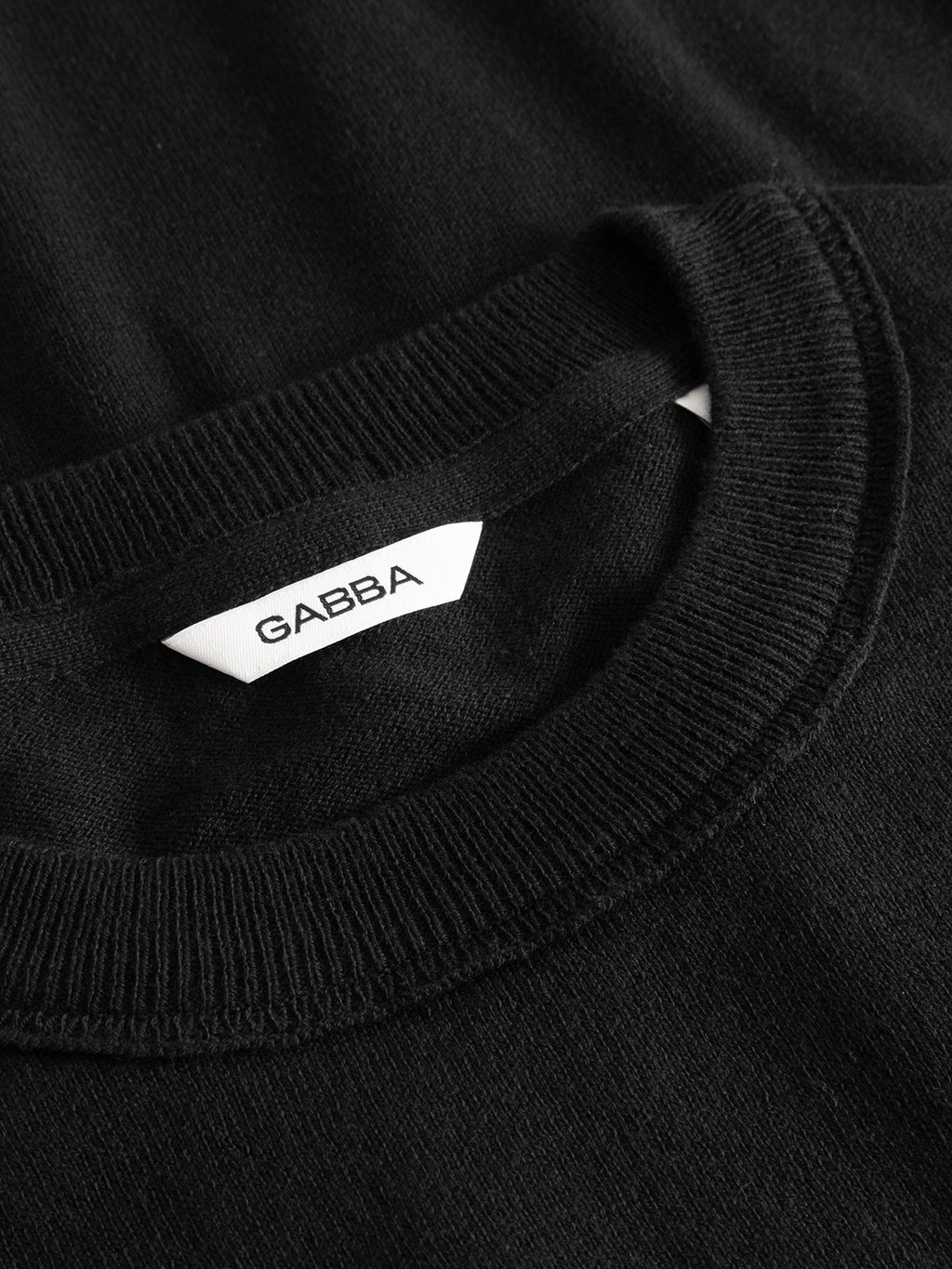 Close-up of the GABfumo Spy LS Knit - Black by Gabba, a crewneck T-shirt with a ribbed, flat-knit collar and a white tag showing the Gabba brand name in black letters.