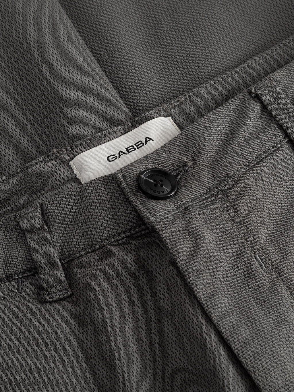 Close-up of the waistband of Gabba’s GABpaul K3280 Dale Chino in Dust Sage Green, featuring a black button, belt loop, white label, subtly textured fabric, and classic French pockets for stylish detail.