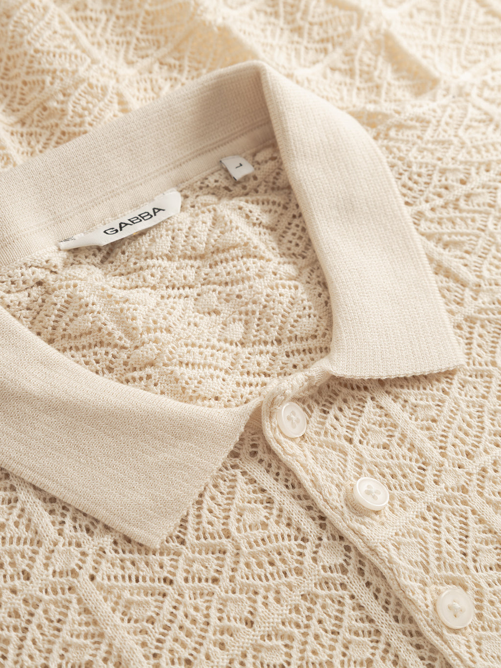 Close-up of the Gabba GABbenson Fiwi SS polo in Birch—a cream-colored short-sleeve knit with a textured geometric pattern, ribbed collar, and three white buttons. Brand label visible inside the collar.