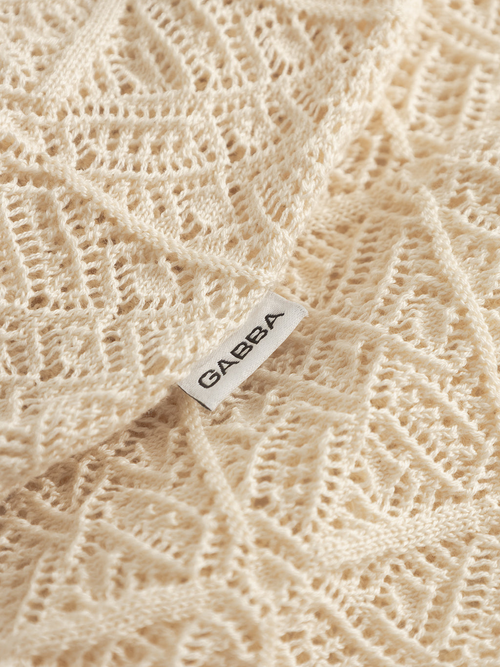 A close-up of cream-colored knit fabric from the Gabba GABbenson Fiwi SS - Birch, featuring intricate open hand-knit patterns and a white Gabba label sewn on.