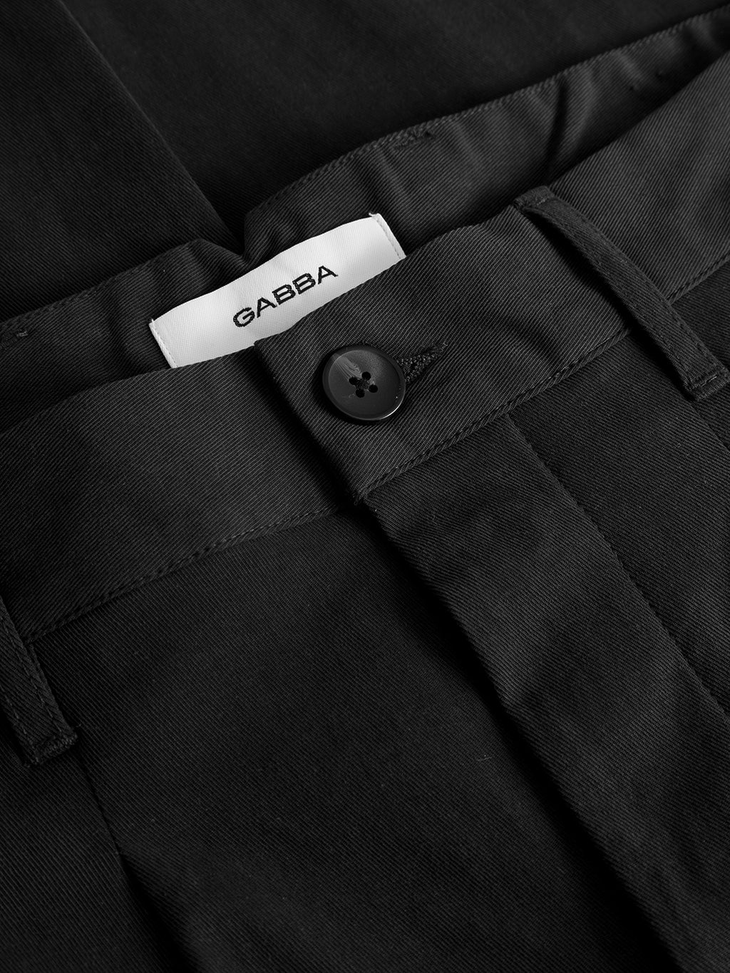 Close-up of the GABfirenze Bolu Cropped TENCEL™ Pant in black by Gabba, featuring button closure and belt loops. A white label inside the waistband displays the Gabba brand. Crafted from smooth, stretch TENCEL™ twill, these pants offer a relaxed fit with a cropped length for contemporary style.