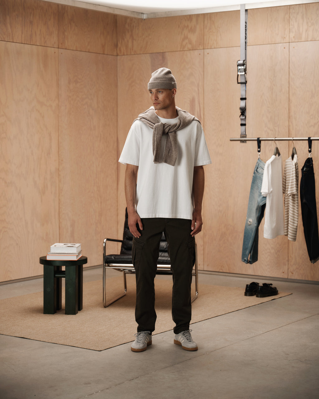 A man wearing a beanie, the Gabba GABnigel Wix Boxy Hour SS t-shirt in white, a sweater draped over his shoulders, dark pants, and sneakers stands in a minimalist room featuring hanging clothes, a chair, a mat, and a small table with books.