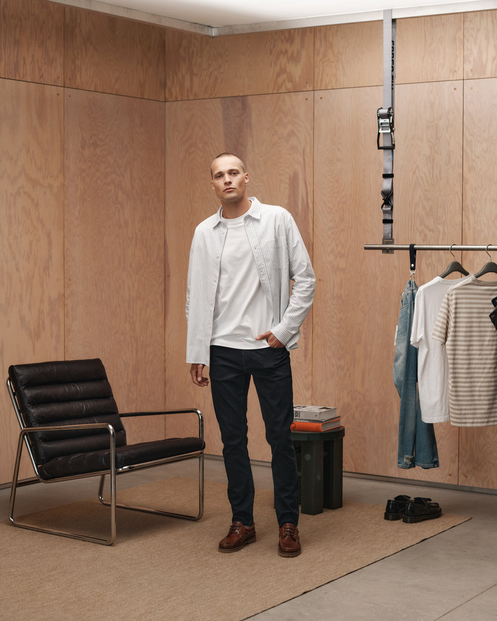 A person stands in a minimalist room with light wood walls, near a black chair, clothes rack with casual wear, and stacked books. They wear a white shirt layered over a white T-shirt, paired with Gabba’s GABjones BlueBlack - Blue Black Denim slim straight fit pants and brown shoes.