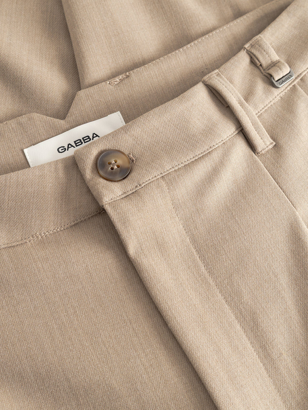 Close-up of GABfirenze Phin Cropped GRS trousers by Gabba in Sand Plaza: beige fabric, brown button, visible belt loops, and a white Gabba label partly tucked into the waistband.