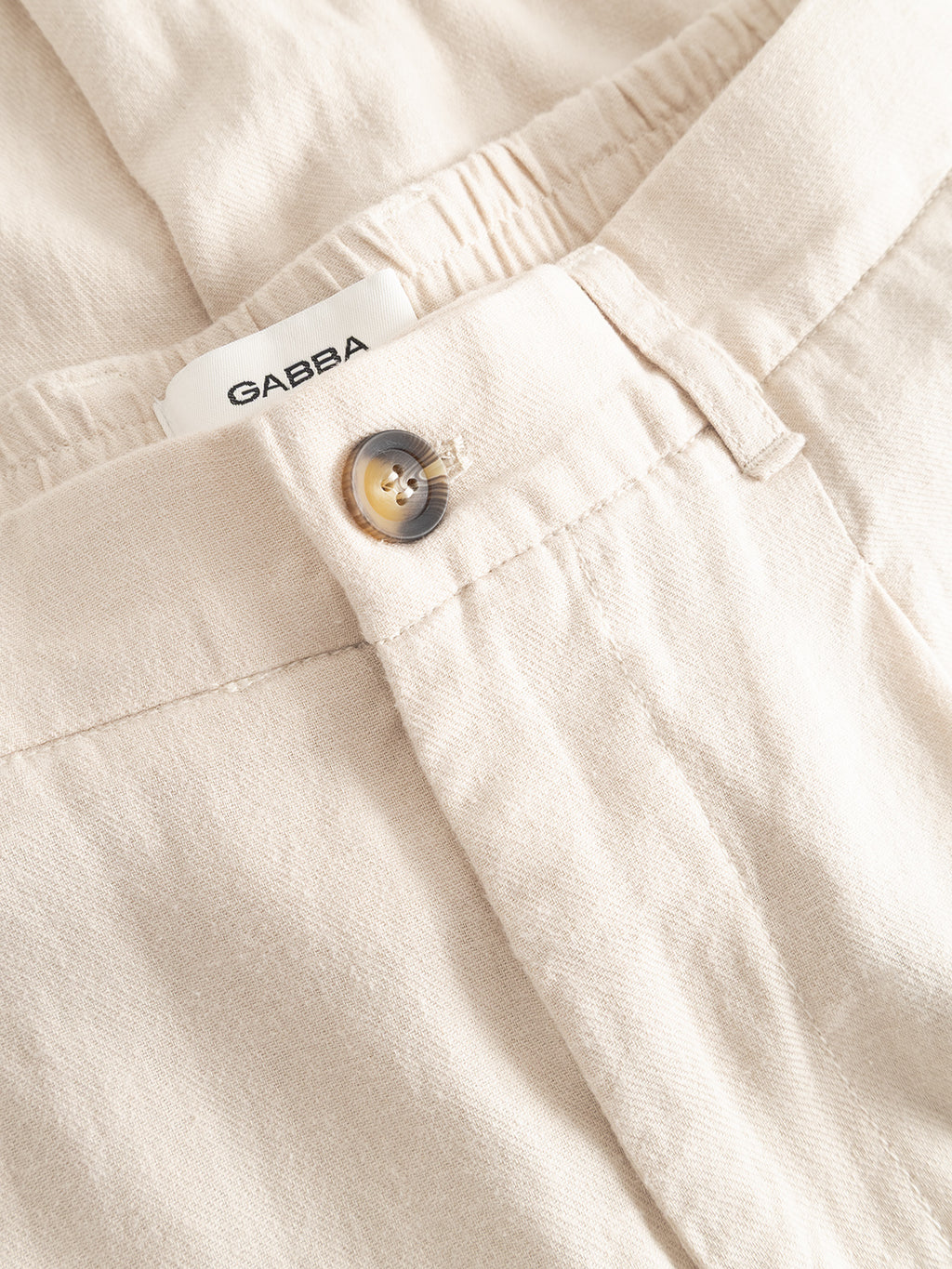 Close-up of Gabba GABnapoli 19 Lino Pant in Birch: beige tailored chinos with front button, elastic waistband, visible white Gabba label, lightweight soft-textured fabric, neat stitching, and a comfortable straight fit.