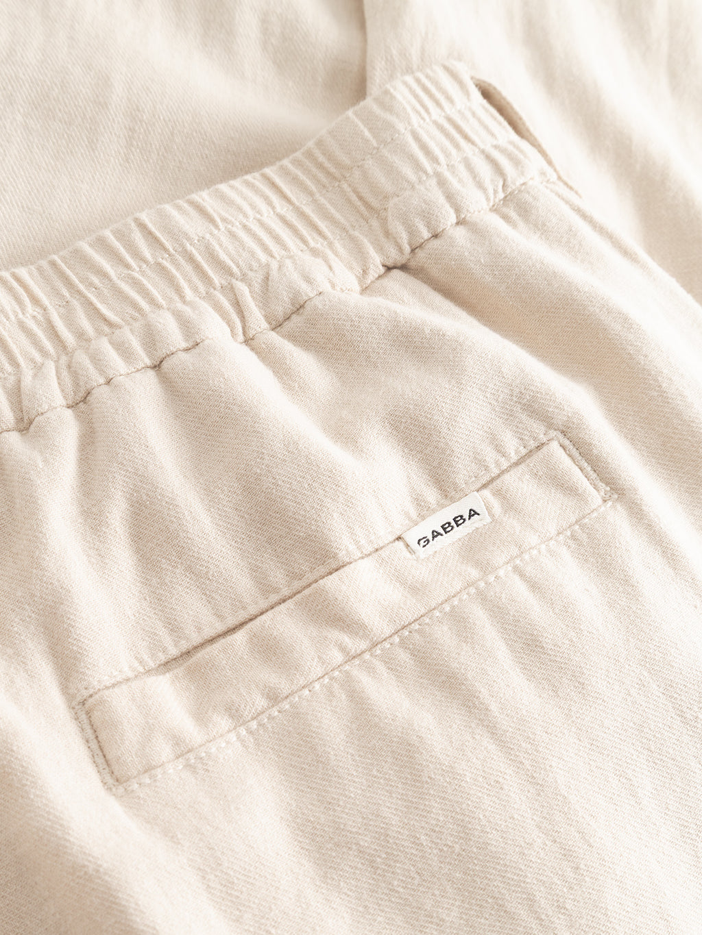 Close-up of the Gabba GABnapoli 19 Lino Pant in Birch, made from lightweight fabric with a regular straight fit, elastic waistband, back pocket, and a small white BAEBA tag above the pocket.