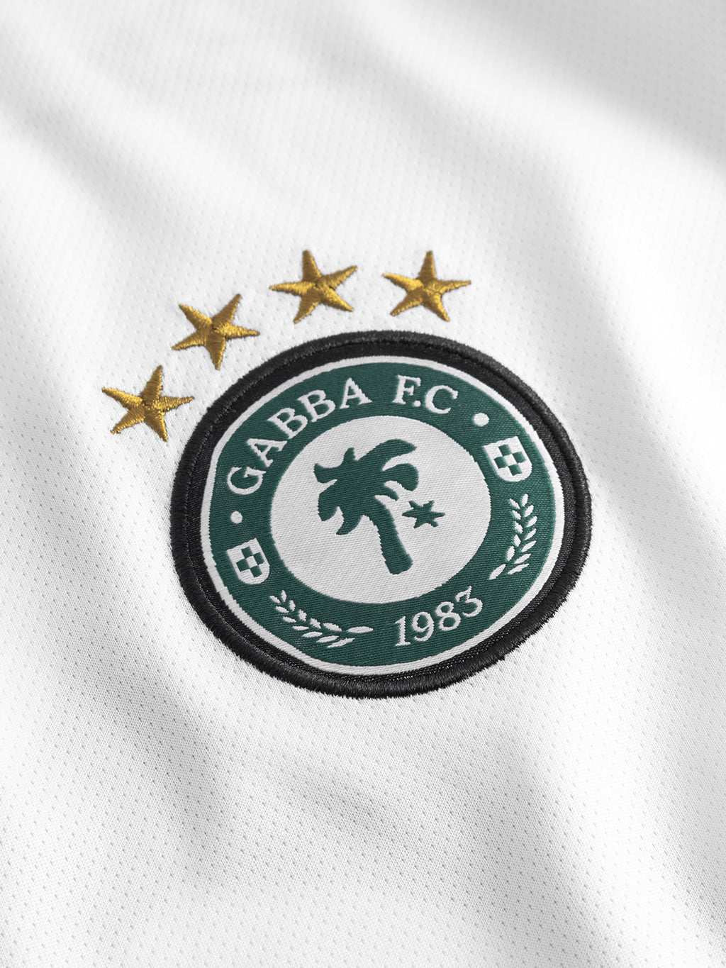 The Gabba GABnigel Fut Boxy Pes SS T-shirt in white reflects football culture with a circular GABBA F.C. 1983 logo, a palm tree center, and four gold stars atop the crest.
