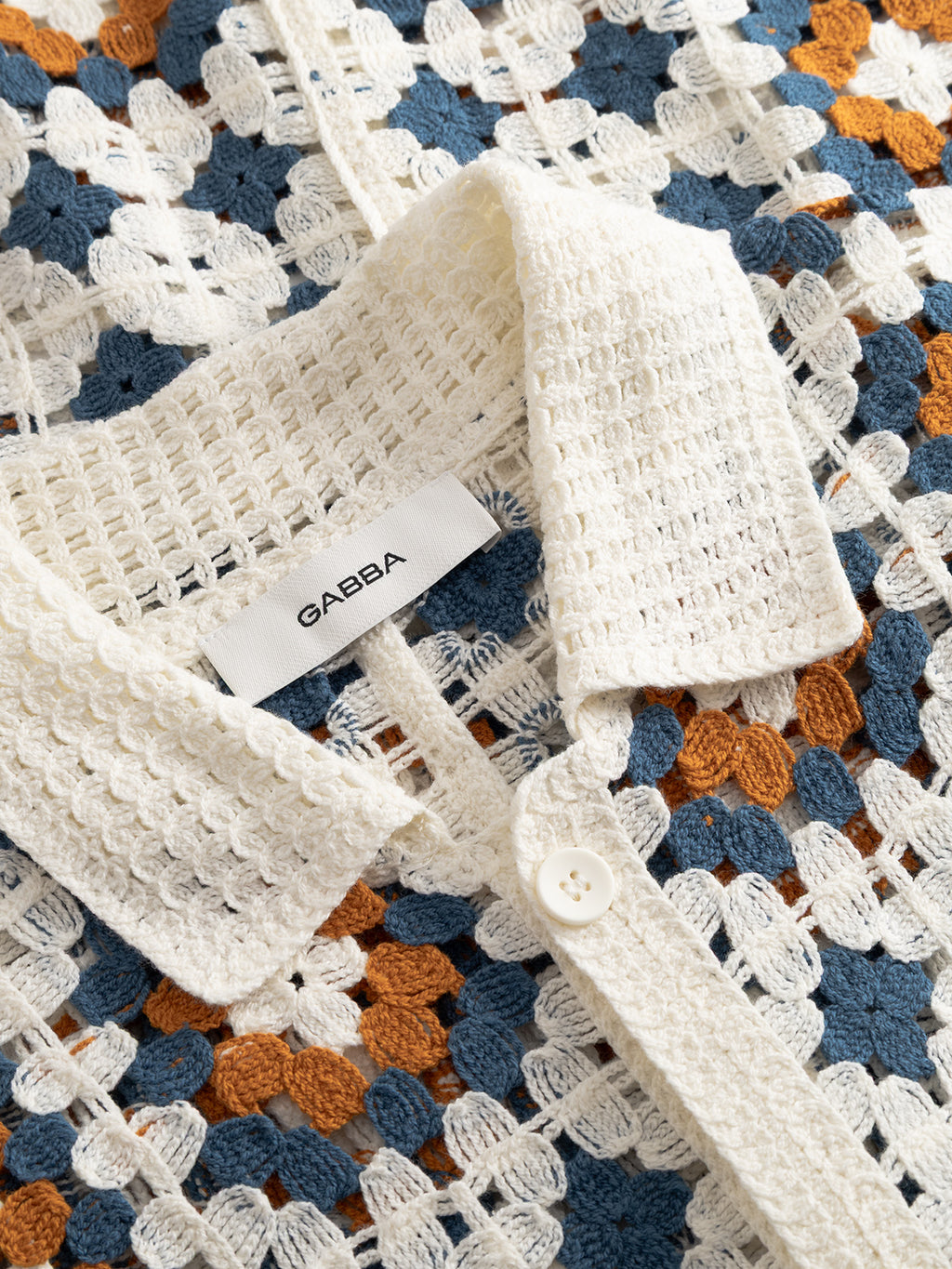 Close-up of the Gabba GABcrochet Pattern SS Knit - Multi Pattern shirt, featuring a white collar, button, and brand label. The open-knit design combines white, blue, and brown yarns for stylish comfort—ideal for layering.