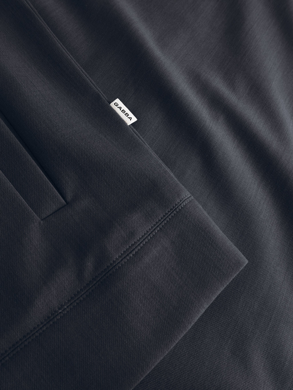 Close-up of the GABnik Phin GRS - Blue Black jacket by Gabba, highlighting a pocket seam and a small white BARDA tag. The smooth, finely textured fabric is made from recycled polyester.