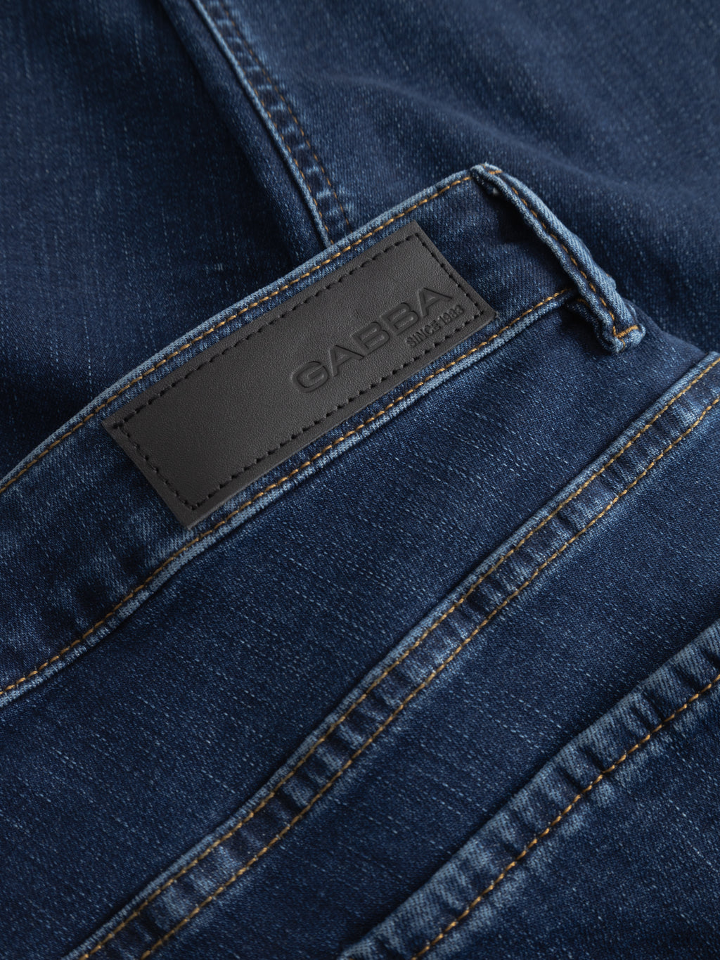 Close-up of GABjones Dhera Jeans in mid blue denim by Gabba, highlighting the back pocket and a black leather patch with the Gabba brand name stitched on. These slim straight fit jeans feature visible orange stitching and a distinctive diagonal seam above the pocket.