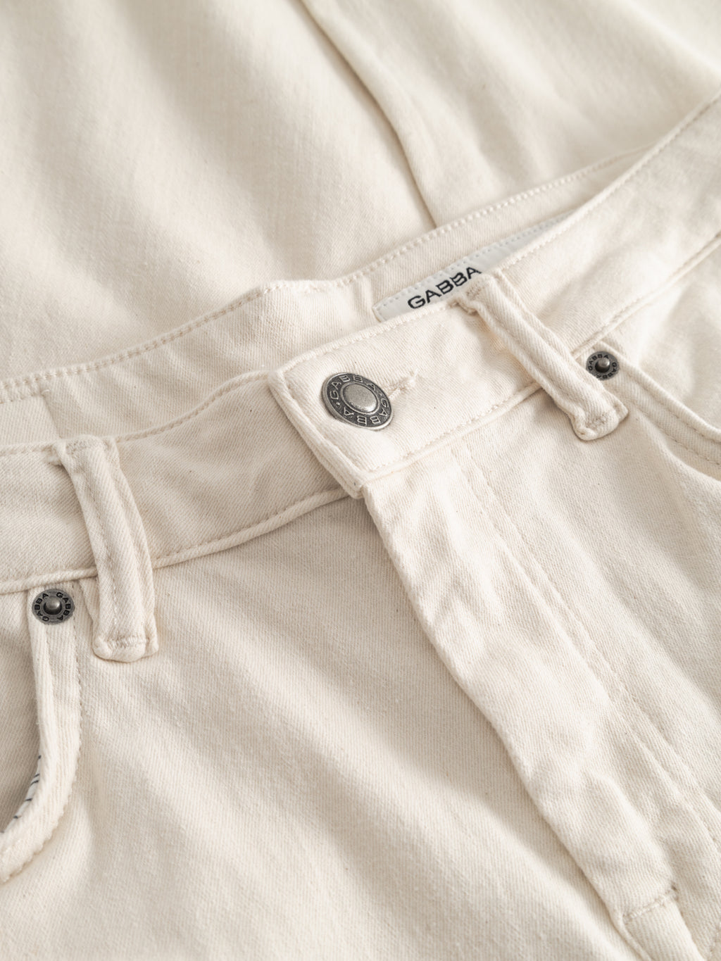 Close-up of the waistband and upper front of GABjones Heski Jeans - Ecru by Gabba, featuring a metal button, belt loops, superstretch fabric, and a visible Gabba label above the zipper.