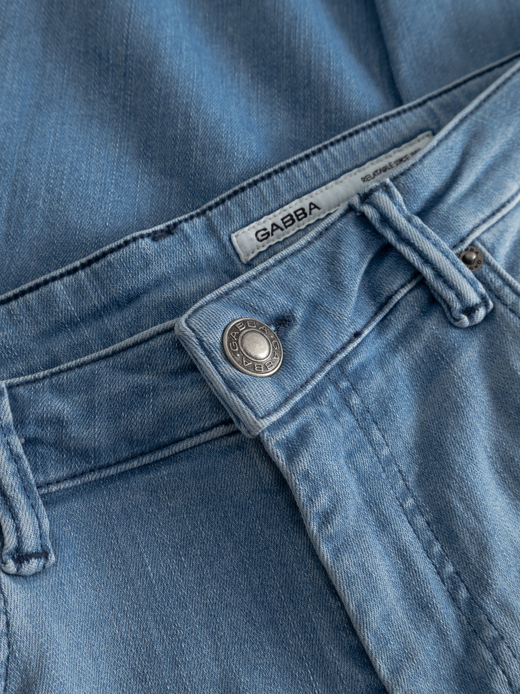 Close-up of GABjones Mik Jeans in light blue denim by Gabba, slim-fit with metal button, belt loops, seam details, and a white fabric brand label, crafted from premium denim.