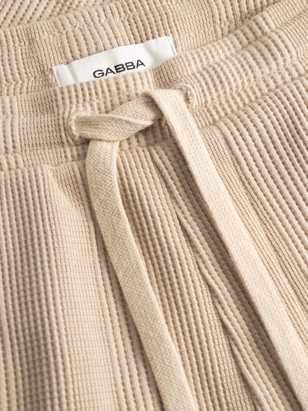 Close-up of GABtommi Wup Shorts - Humus by Gabba in beige waffle-textured fabric, featuring an adjustable drawstring, elastic waistband, and a white Gabba label sewn inside.