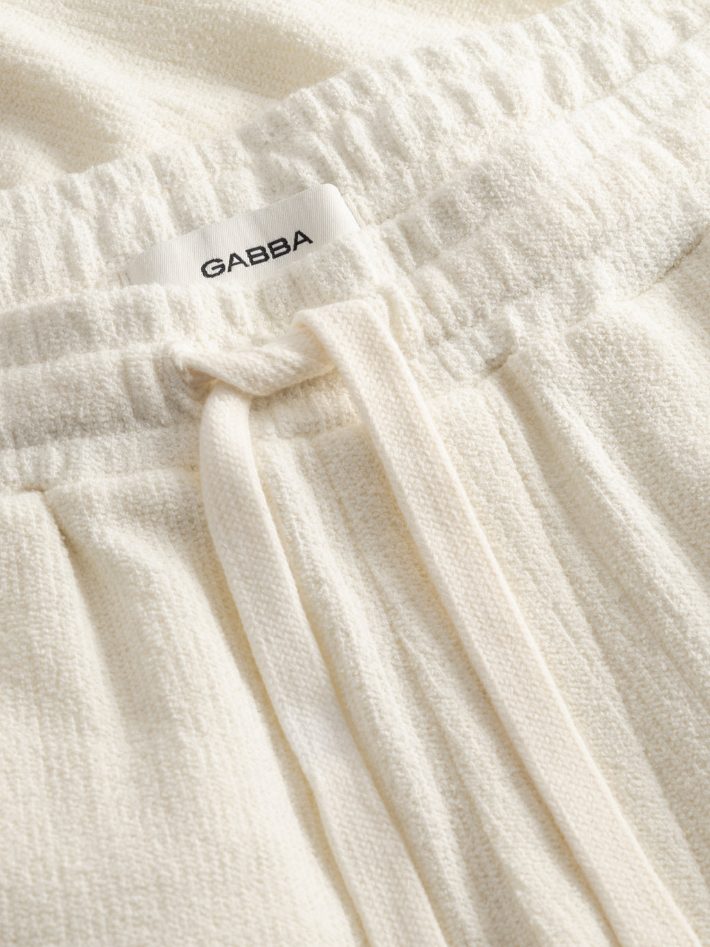 Close-up of GABtommi Towel Shorts in Egret by Gabba, crafted from cream terry cotton with an elastic waistband, drawstring, and a white Gabba label.