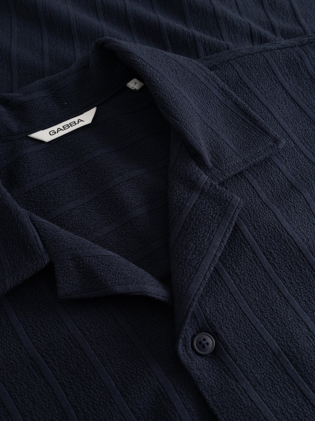 Close-up of the Gabba GABvalente Armor SS T-Shirt in navy, featuring a textured cotton-blend, short sleeves, vertical stripes, a resort collar, a partially visible button, and an inner tag labeled Gabba.