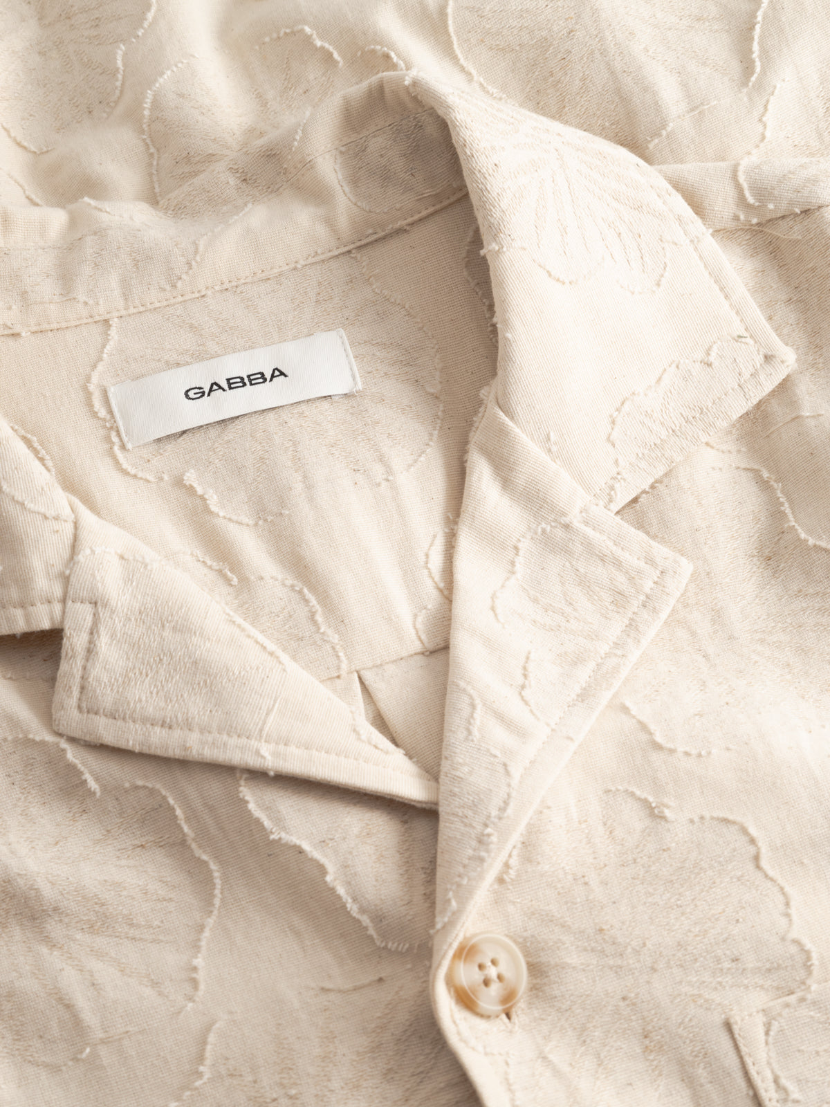 Close-up of the Gabba GABaustin Reso Raw LS Overshirt in Lt. Sand, a cream-colored jacquard-woven cotton shirt with a subtle floral pattern, notched collar, visible button, and inner white Gabba label.