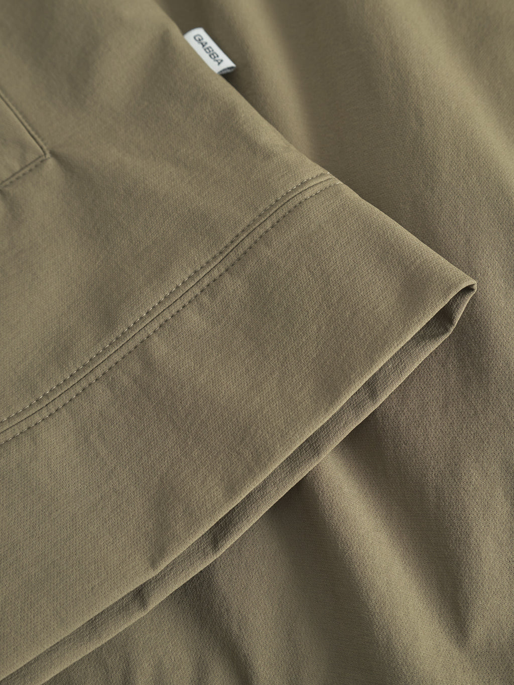 Close-up of the sleeve and hem of the Gabba GABnik Tech - Army, made from durable tech material in olive green, highlighting detailed stitching and a small white fabric tag on the seam.