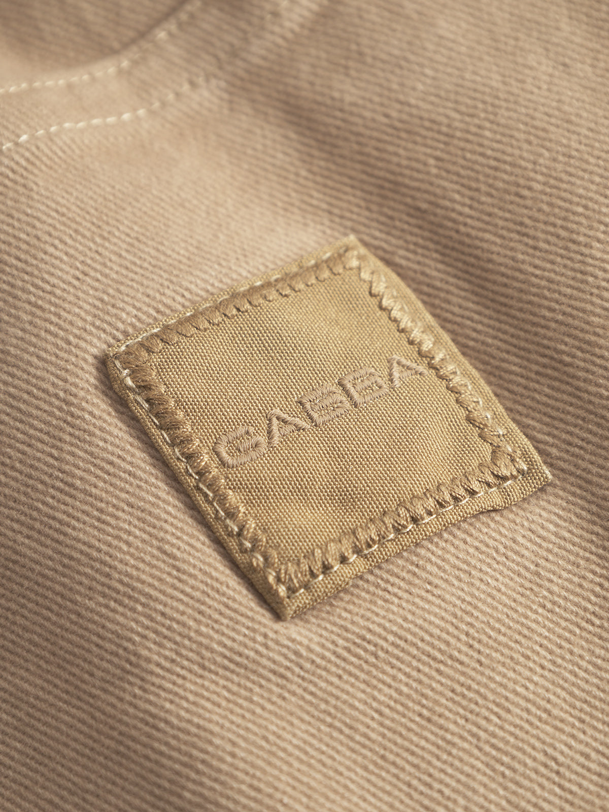 A close-up of a beige GABBA patch with stitched edges, embroidered onto sand-colored GABbeta Twill by Gabba, highlighting the classic workwear-inspired style of this overshirt.