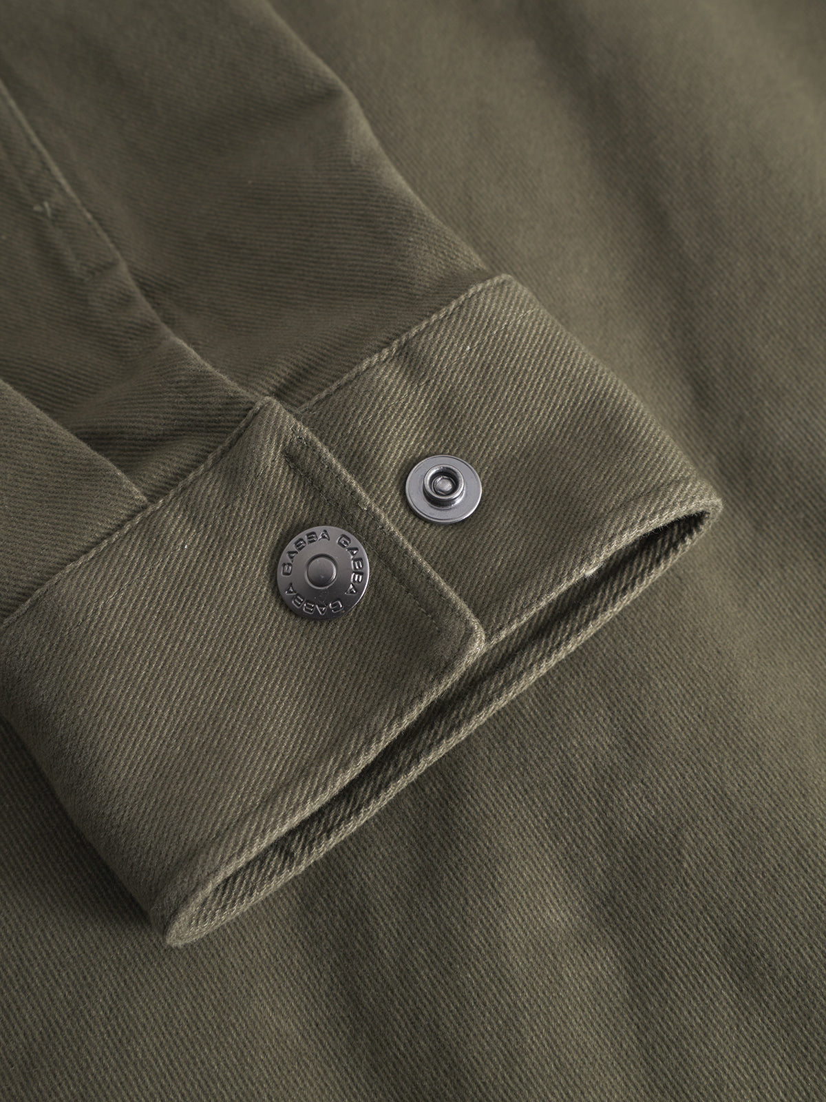 A close-up of an olive green GABbeta Twill - Army cuff by Gabba shows visible stitching, a metal snap button, and a GADEA BILBAO-engraved rivet—ideal for a comfortable shirt or workwear overshirt.