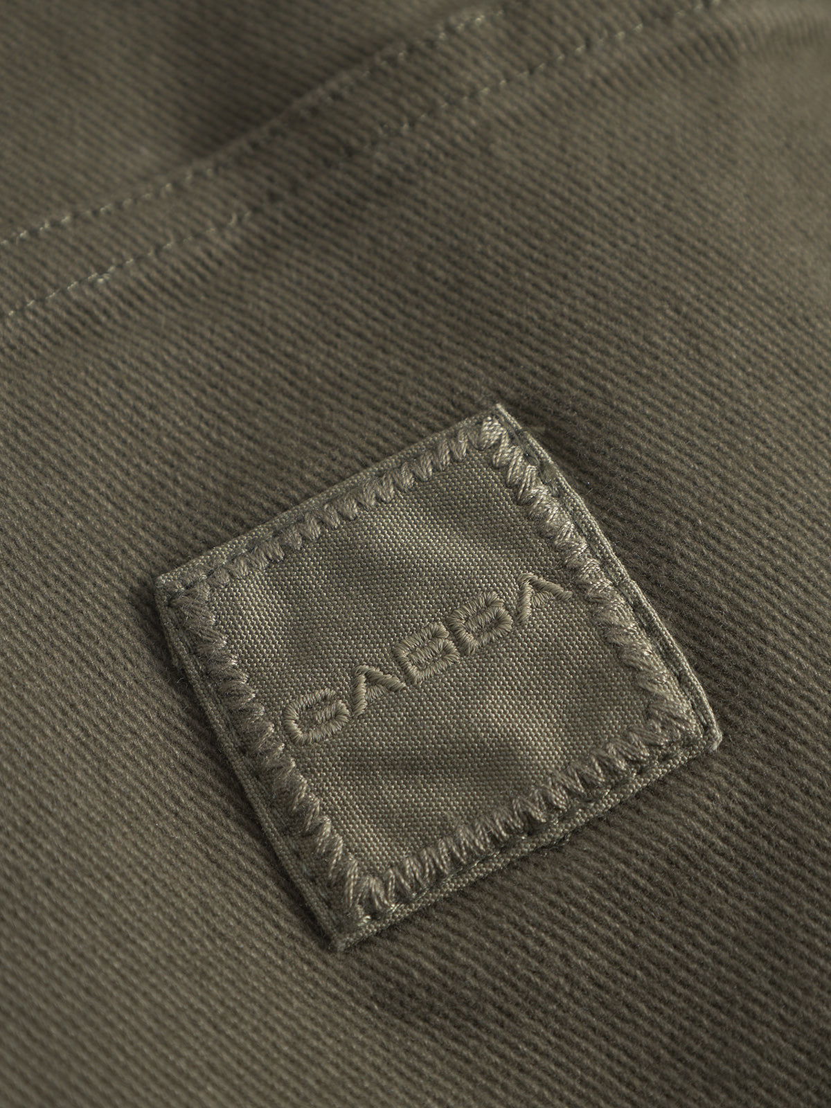 Close-up of the GABbeta Twill - Army by Gabba: khaki garment dyed twill with a stitched square patch labeled GABBA in raised letters. Quality stitching and texture make it ideal as a comfortable workwear overshirt.
