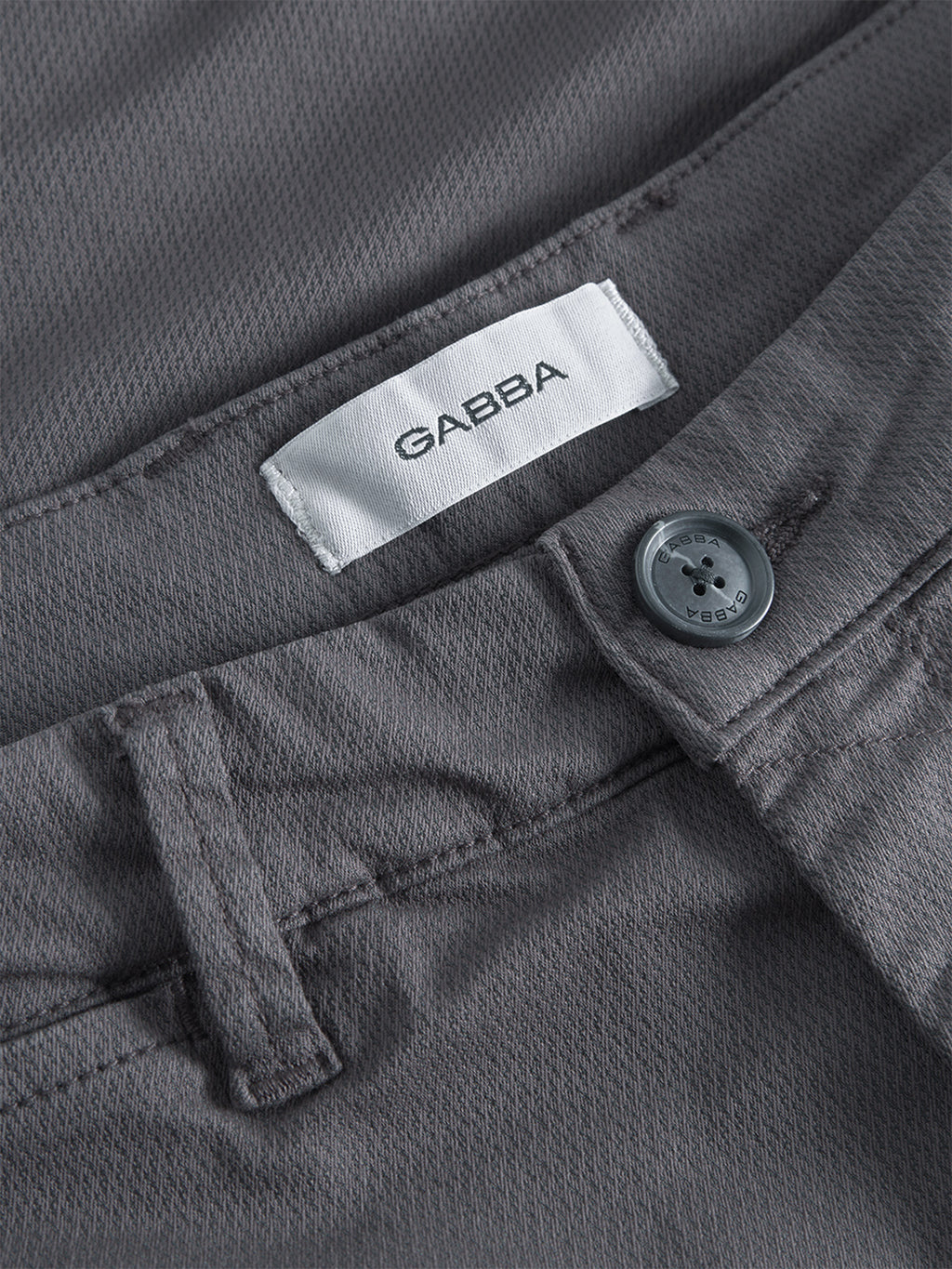 Close-up of GABpaul K3280 Dale Chino in charcoal by Gabba, showcasing textured gray fabric, waistband with belt loops, button, French pockets, and a white branded label for a classic style.