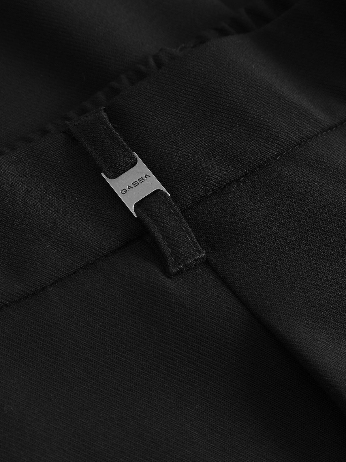 Close-up of the Gabba GABnapoli 19 Phin GRS - Black pants, featuring a silver metal tag labeled Gabba attached to a belt loop. The texture of the recycled polyester fabric and detailed stitching are visible.