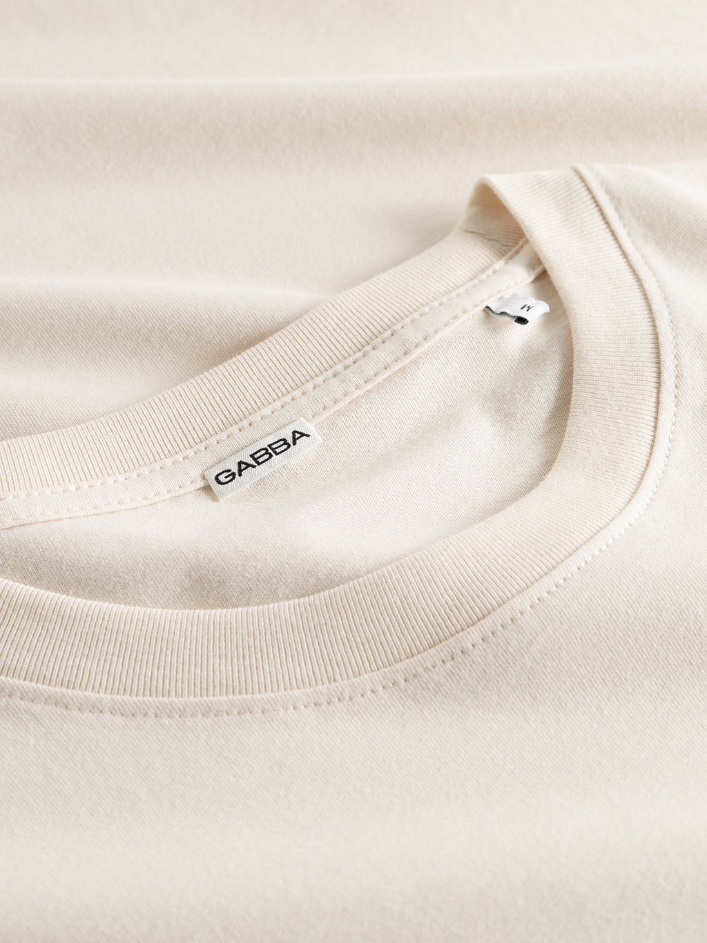Close-up of the GABdune Logo GOTS SS T-Shirt in Birch by Gabba, made from light beige organic cotton, laid flat to display its crew neckline and a white Gabba label sewn inside the collar.