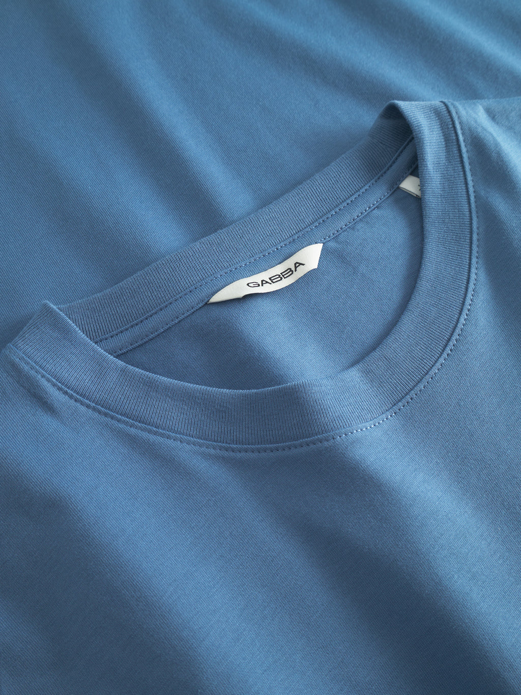 Close-up of a blue Gabba GABdune Logo GOTS SS T-Shirt in Blue Horizon with visible neck label, laid on another blue garment of similar shade.