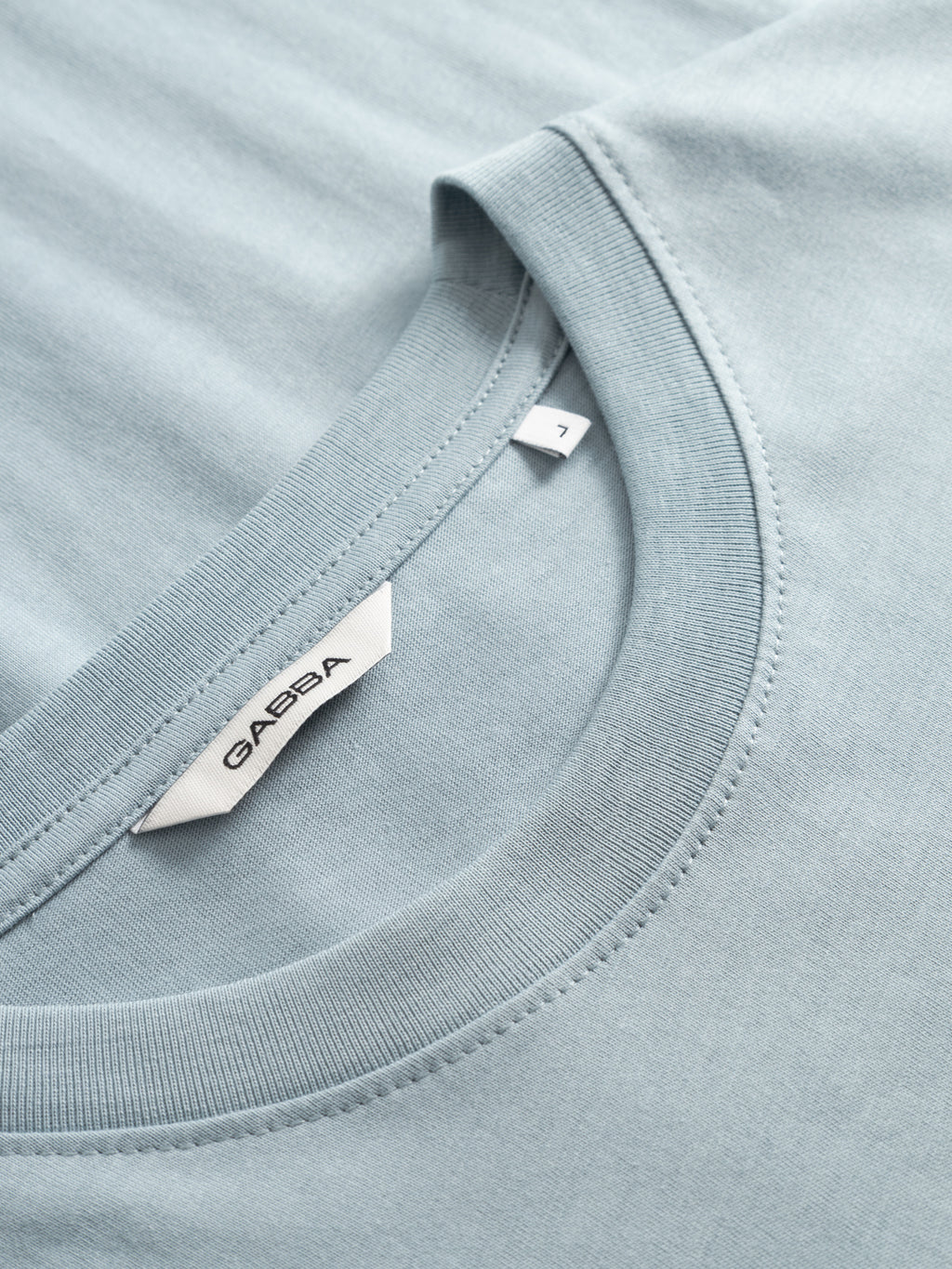 Close-up of a light blue Gabba GABdune Logo GOTS SS T-Shirt, neatly folded. The soft organic cotton fabric is shown with the Gabba label inside the neck and a small white tag marked size 7 near the collar.