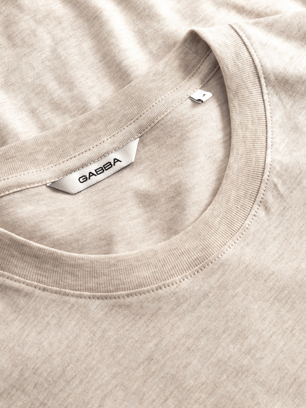 A close-up of the GABdune Melange New SS T-Shirt in Sand Mel. by Gabba, featuring soft beige melange jersey fabric and a white label with the brand name inside the collar. The shirt is neatly laid out.