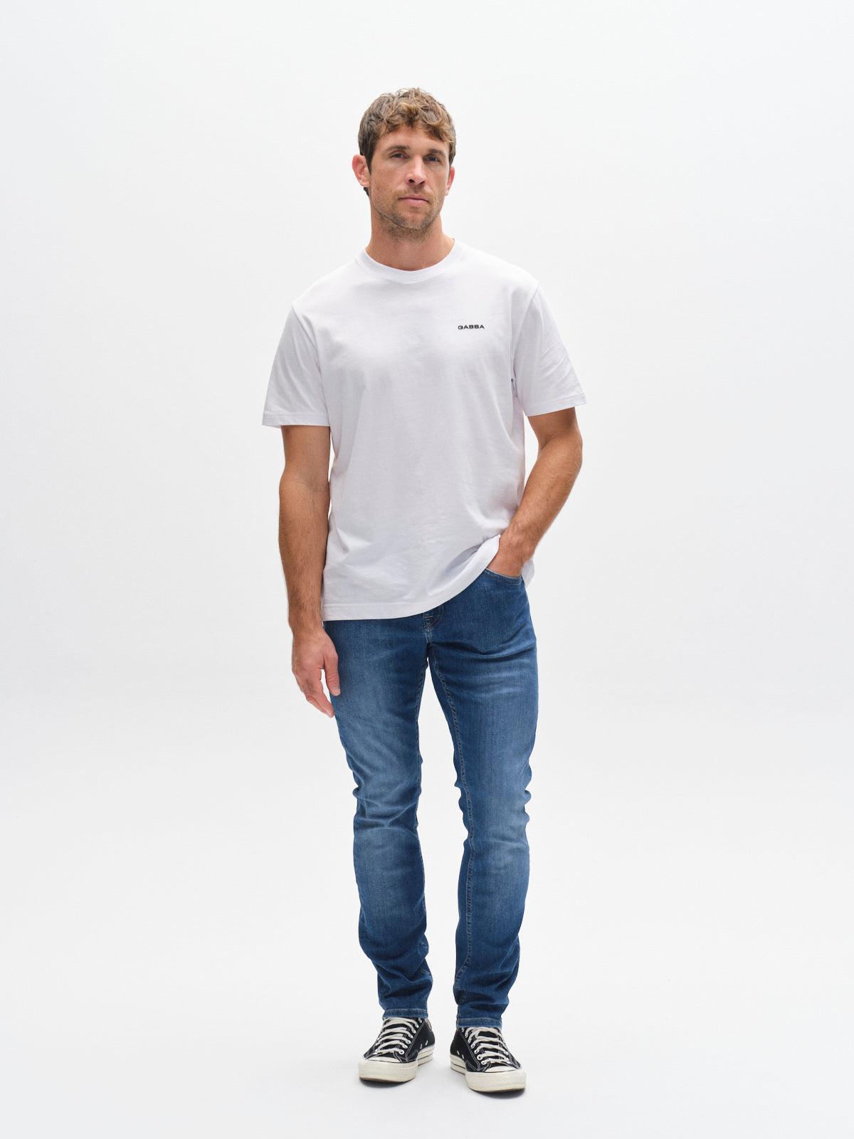 A man models Gabba’s GABjones K3870 - Mid Blue Denim jeans with a plain white t-shirt and black sneakers, standing against a white background. With one hand in his pocket, he highlights the stylish versatility of Gabba denim.
