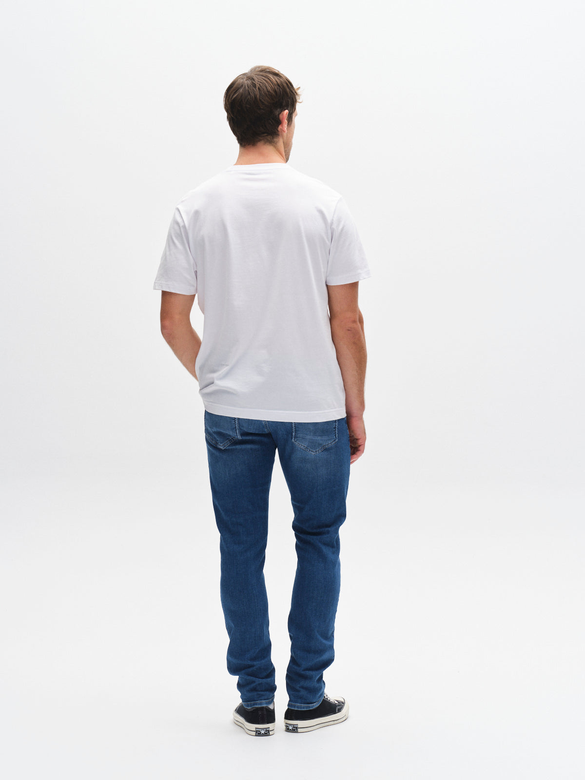 A man stands with his back to the camera against a plain white background, wearing a white t-shirt, Gabba GABjones K3870 - Mid Blue Denim slim straight jeans, and black sneakers.