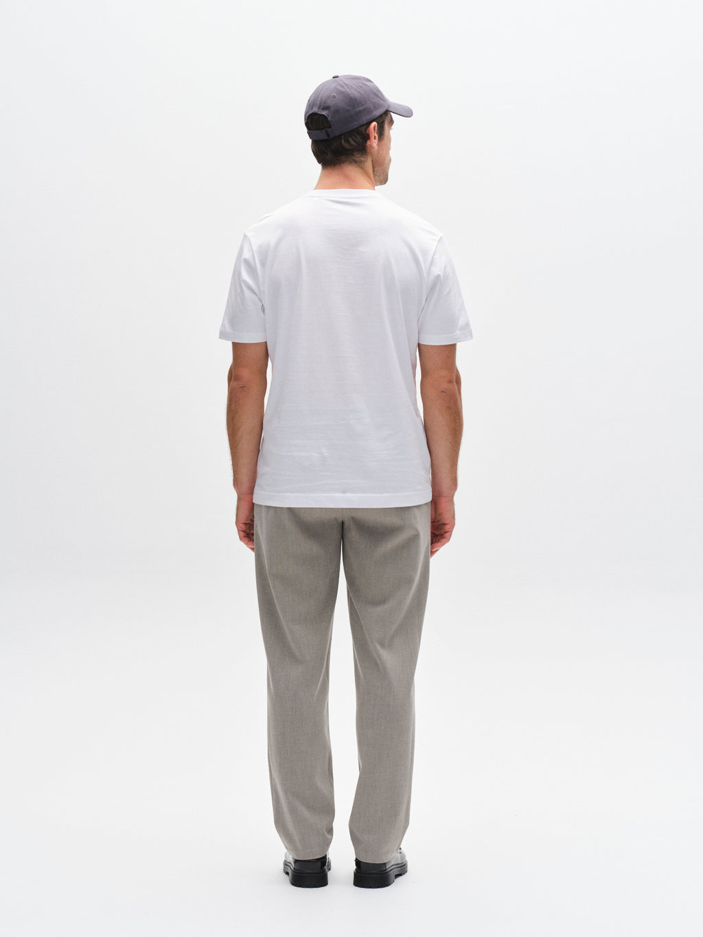 A man stands facing away from the camera, wearing the Gabba GABdune Logo GOTS SS T-Shirt in white, along with light gray pants, black shoes, and a dark gray baseball cap, set against a plain white background.