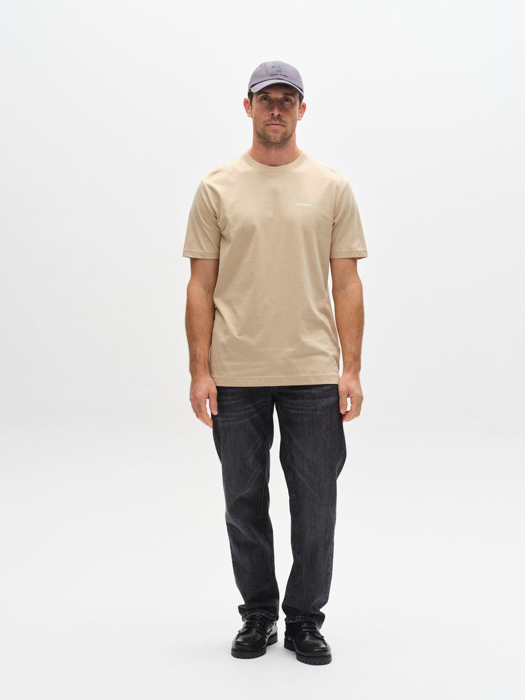 A man stands facing forward against a white background, wearing a gray cap, Gabba GABdune Logo GOTS SS T-Shirt in Safari, dark jeans, and black shoes. His arms are by his sides and his expression is neutral.