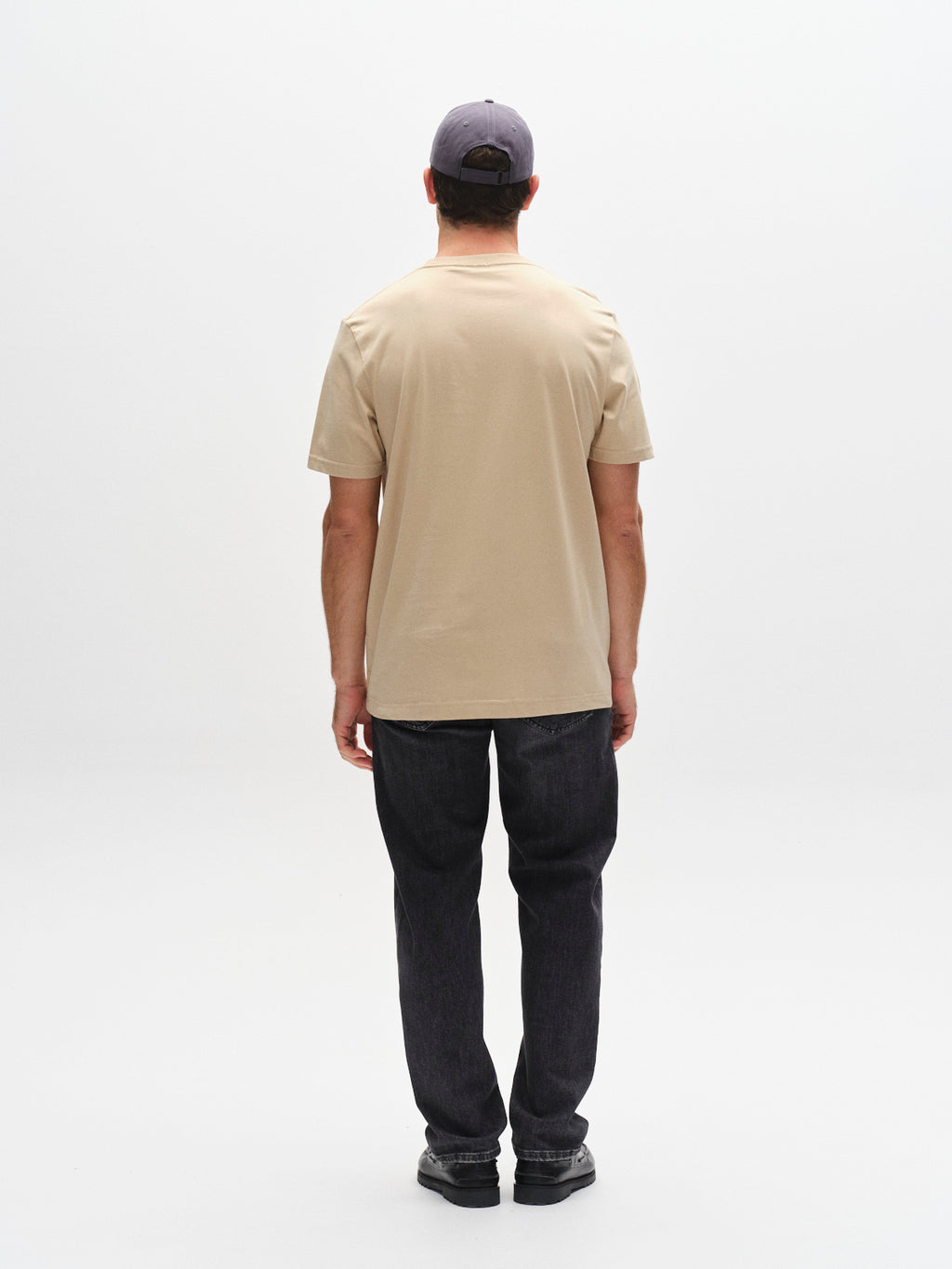 A person faces away from the camera, wearing a Gabba GABdune Logo GOTS SS T-Shirt in Safari, dark jeans, black shoes, and a gray cap against a plain white background.