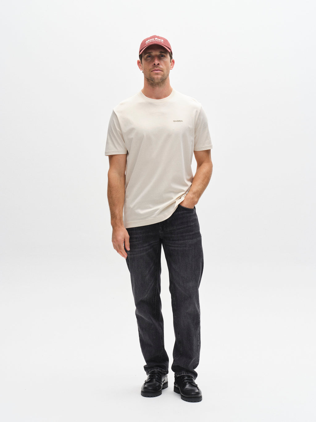 A man stands against a white background wearing the Gabba GABdune Logo GOTS SS T-Shirt in Birch, dark jeans, black shoes, and a red baseball cap. He has one hand in his pocket and a neutral expression.