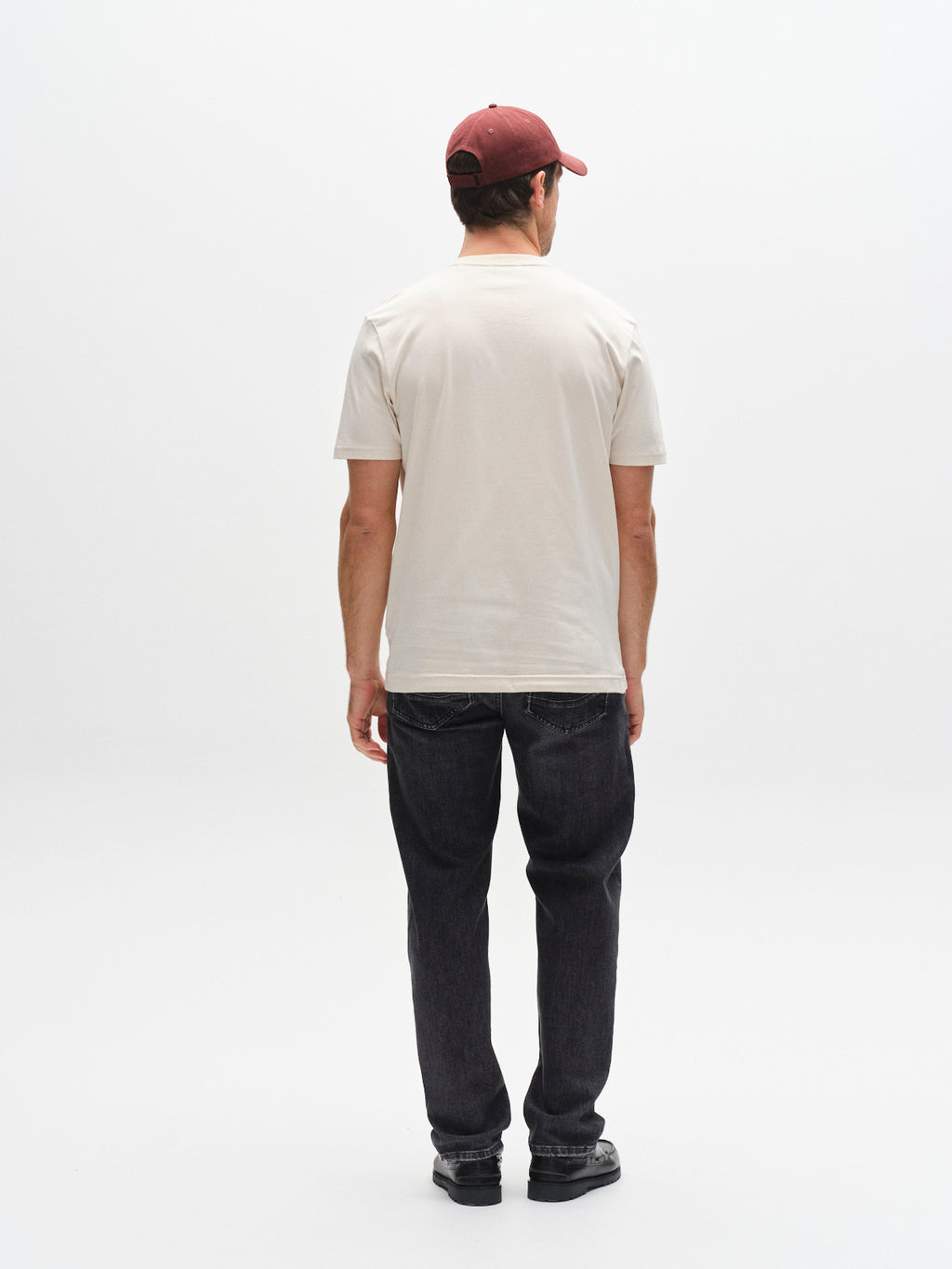 A man faces away from the camera, wearing a Gabba GABdune Logo GOTS SS T-Shirt in Birch, dark jeans, black shoes, and a maroon baseball cap against a plain white background.