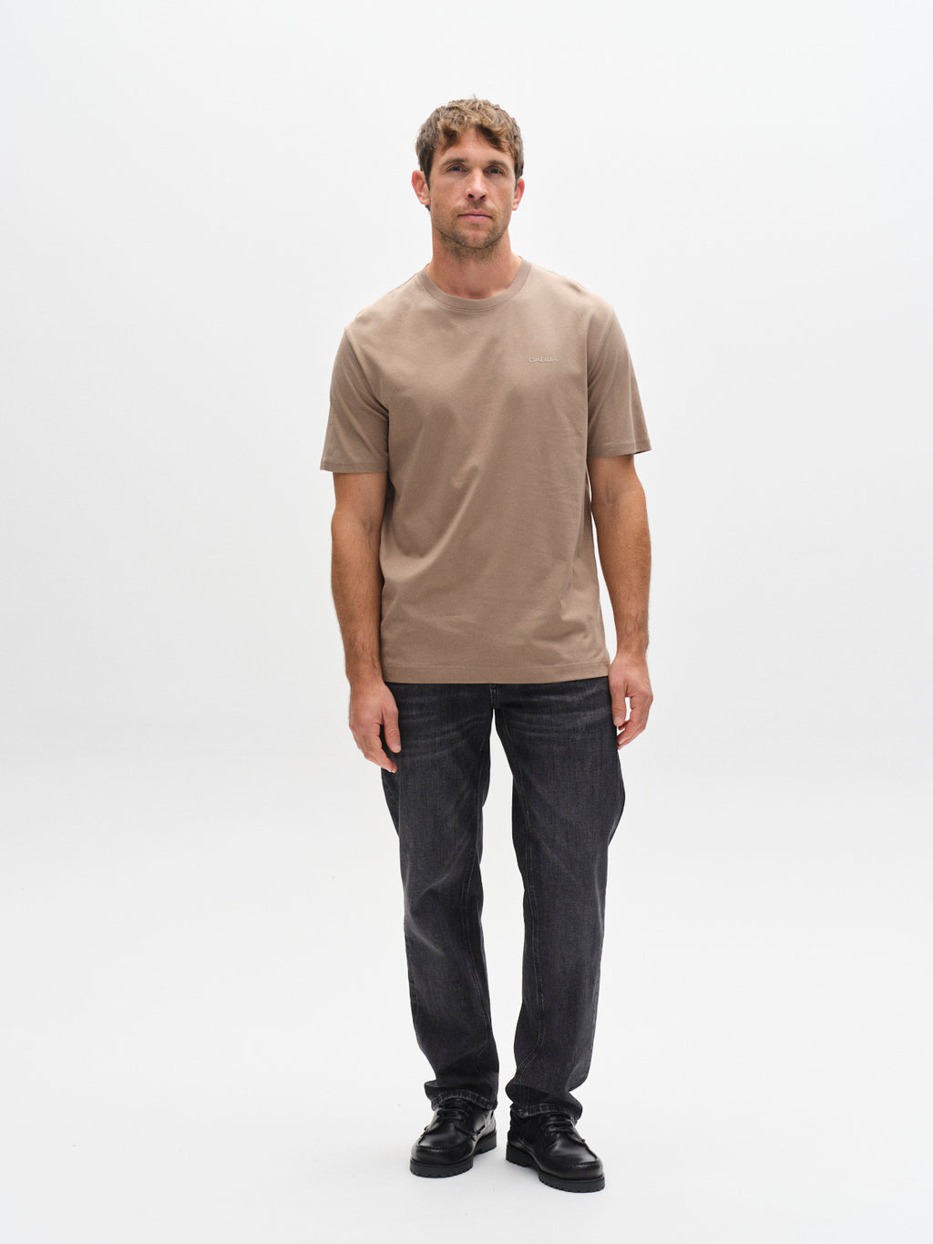 A man with short brown hair stands against a plain white background, wearing the Gabba GABdune Logo GOTS SS T-Shirt in Fungi, dark jeans, and black shoes. He has a neutral expression and his arms are relaxed at his sides.