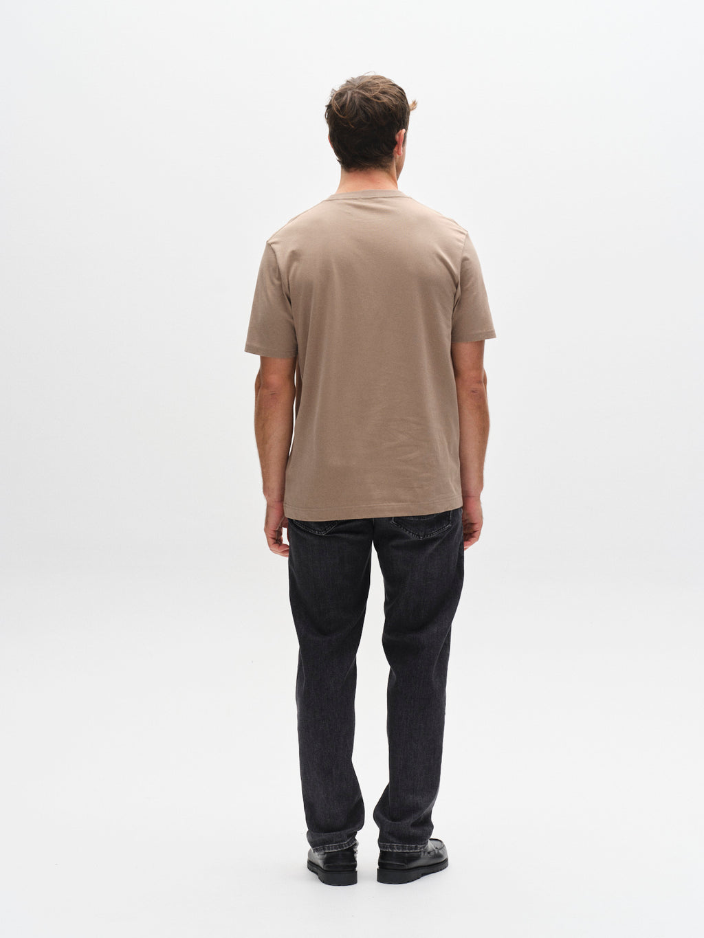 A man faces away from the camera, wearing the Gabba GABdune Logo GOTS SS T-Shirt in Fungi with dark jeans and black shoes, against a plain white background.