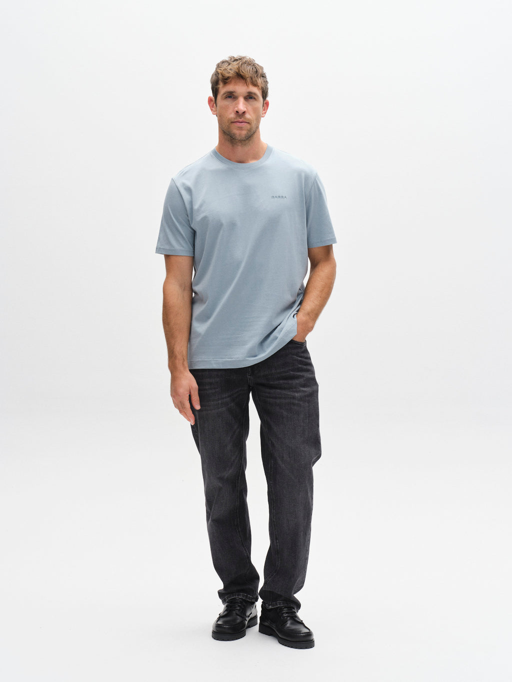 A man stands against a white background, wearing the Gabba GABdune Logo GOTS SS T-Shirt in light blue with dark jeans and black shoes. He has one hand in his pocket and a neutral expression.
