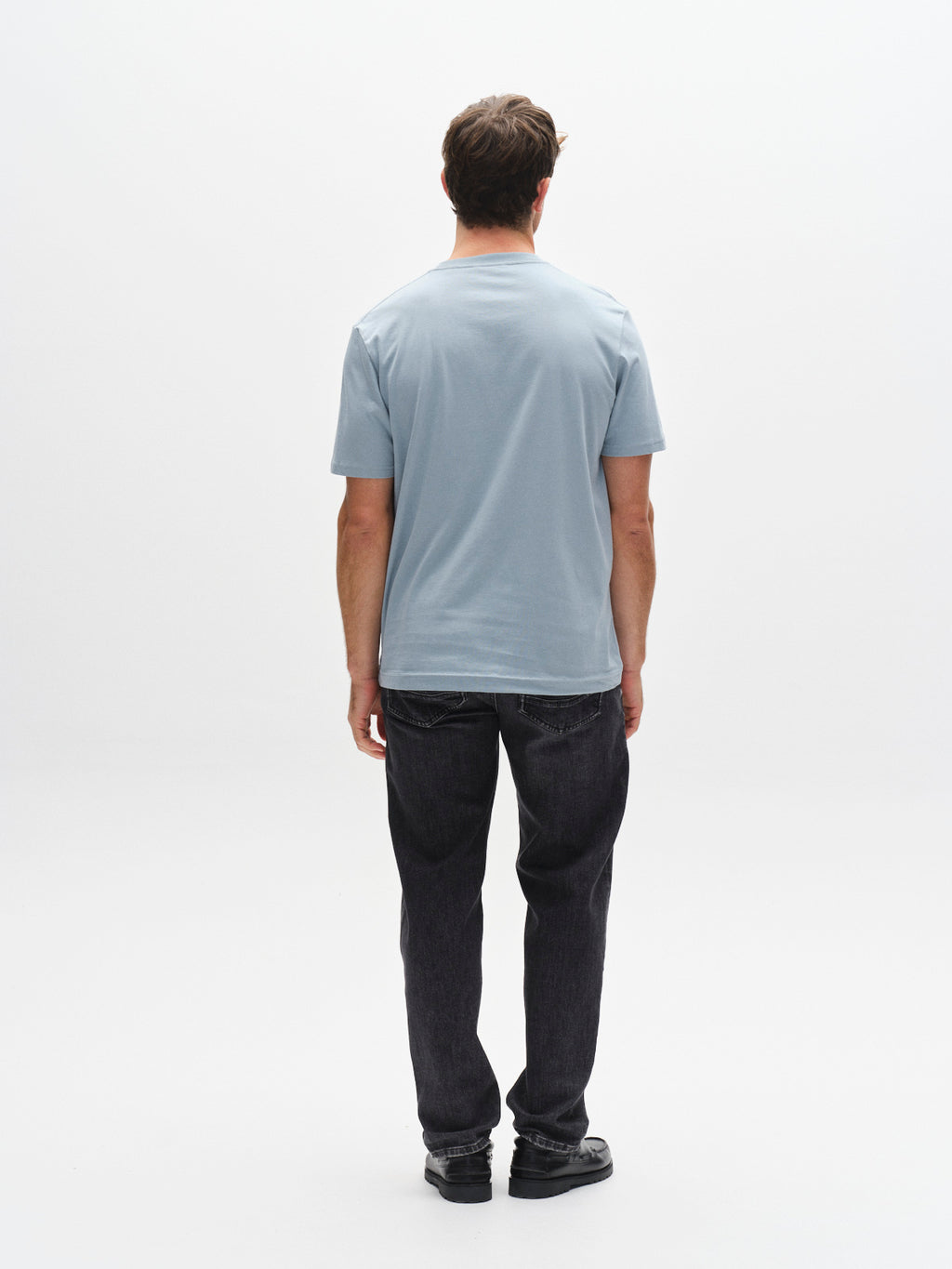 A man stands with his back to the camera, wearing a Gabba GABdune Logo GOTS SS T-Shirt in light blue, paired with dark jeans and black shoes against a plain white background.