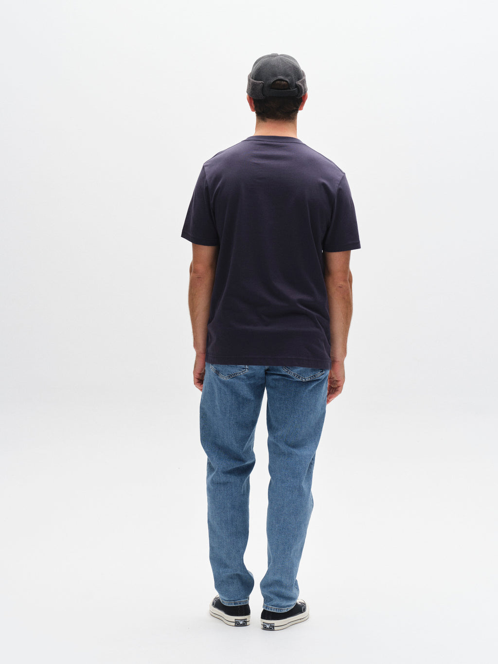 A man faces away from the camera, wearing a Gabba GABdune Logo GOTS SS T-Shirt in Total Eclipse, blue jeans, a black cap, and sneakers against a plain white background.