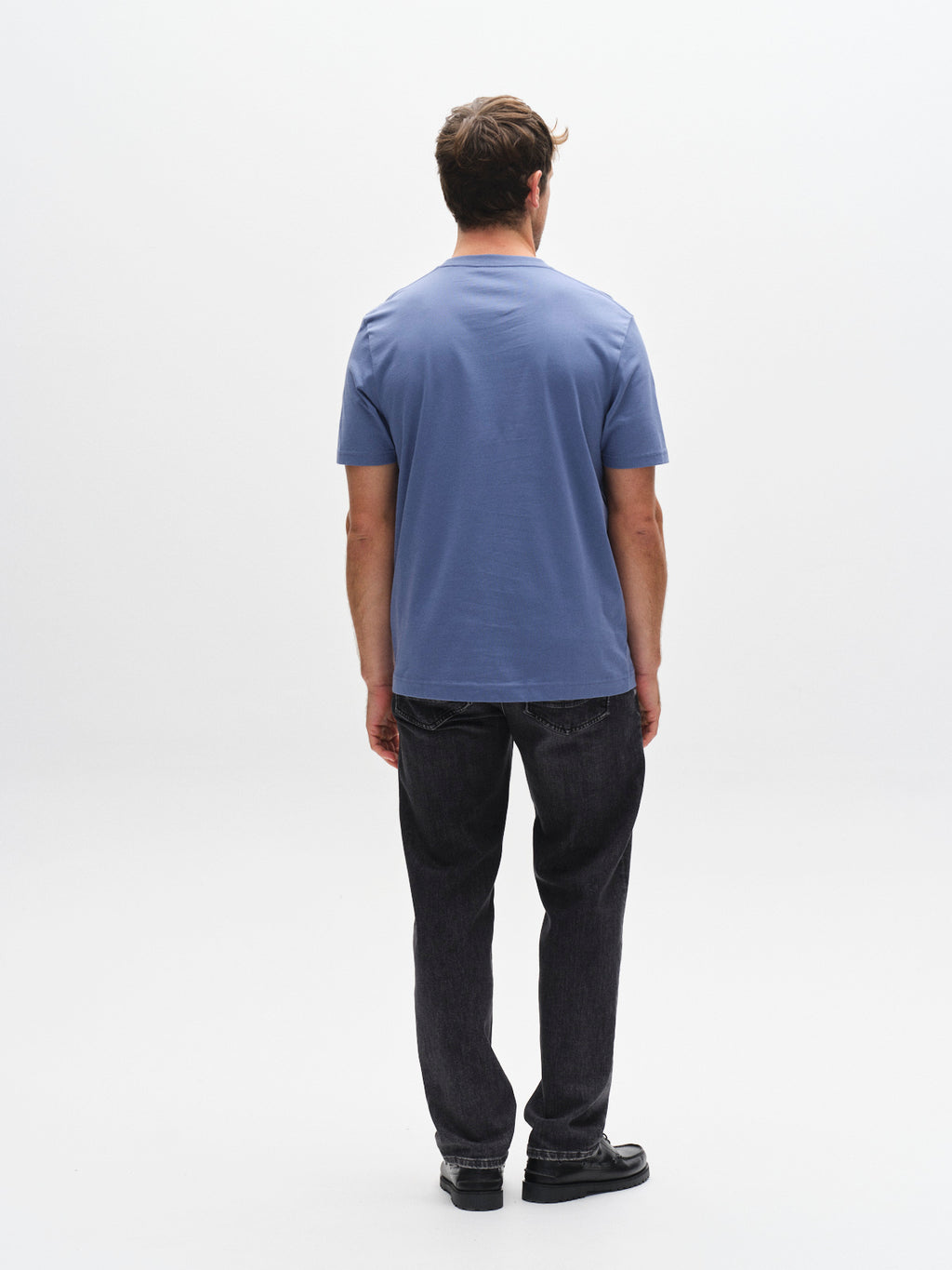 A man stands facing away from the camera, wearing a Gabba GABdune Logo GOTS SS T-Shirt in Blue Horizon, black jeans, and black shoes against a plain white background.