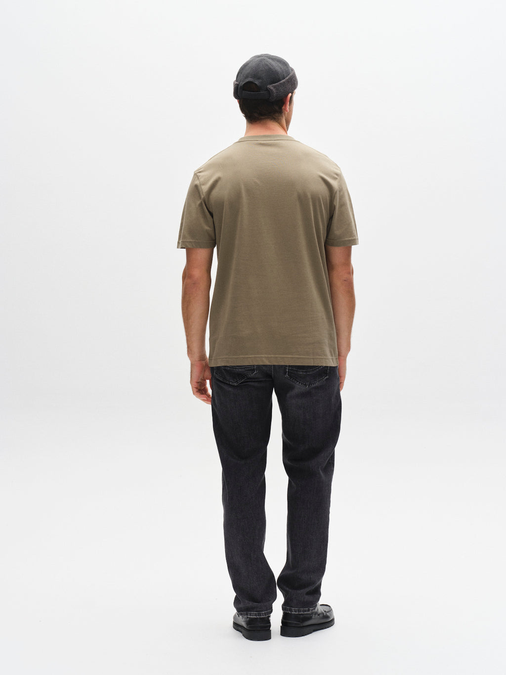 A man faces away from the camera, wearing a Gabba GABdune Logo GOTS SS T-Shirt in Deep Lichen Green, a gray flat cap, dark jeans, and black shoes against a plain white background.